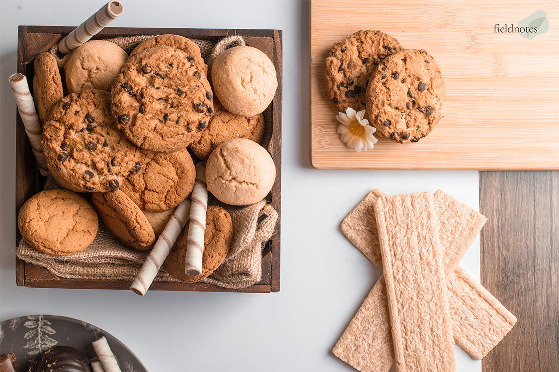 Assorted cookies displayed in a rustic wooden box and on a wooden board, including chocolate chip and plain cookies, with wafer sticks and crackers arranged neatly beside them.