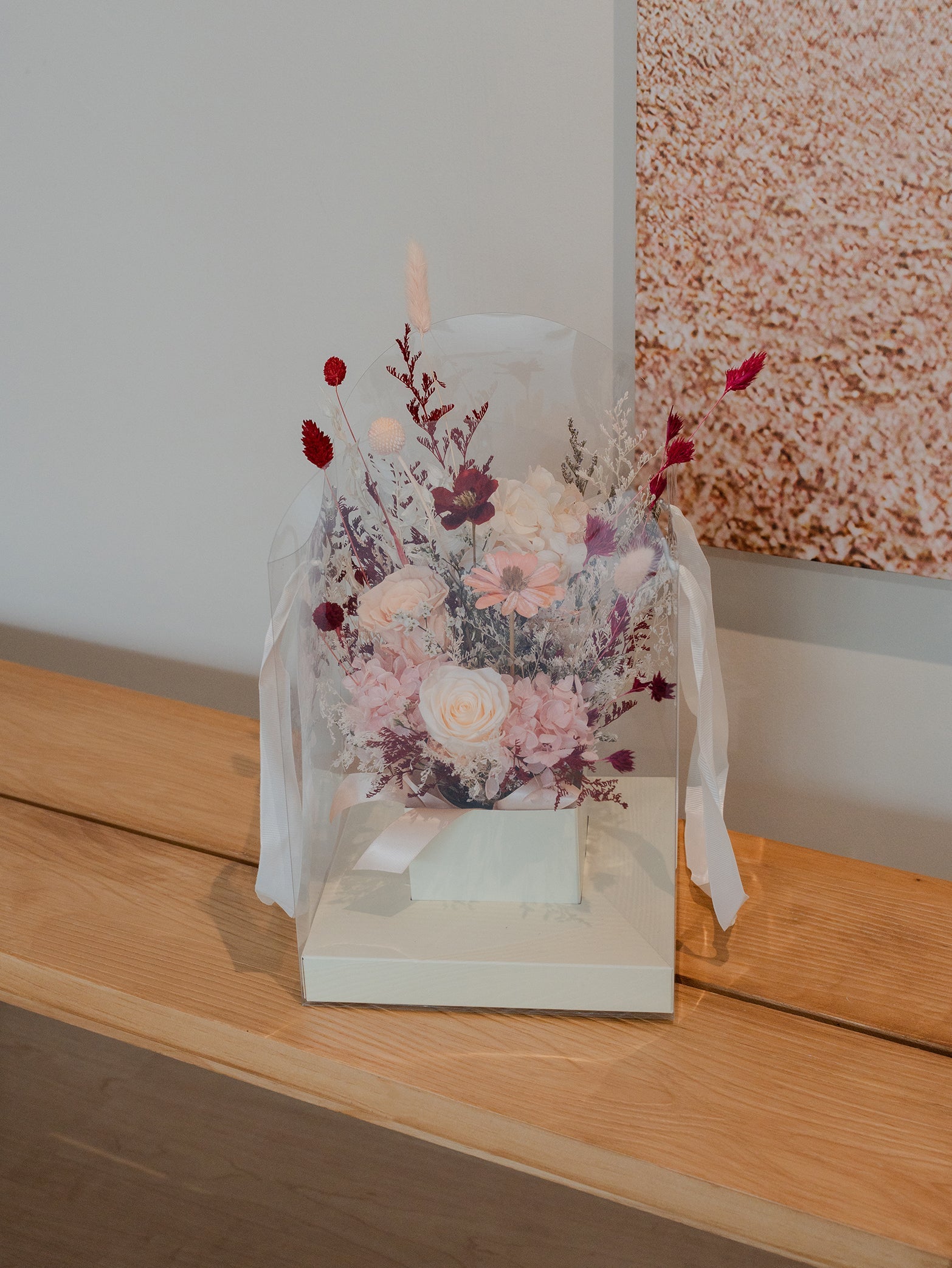 A bouquet of pink, white, and burgundy dried flowers is arranged in a white gift box and wrapped with sheer white ribbon, displayed on a wooden surface next to a framed picture.