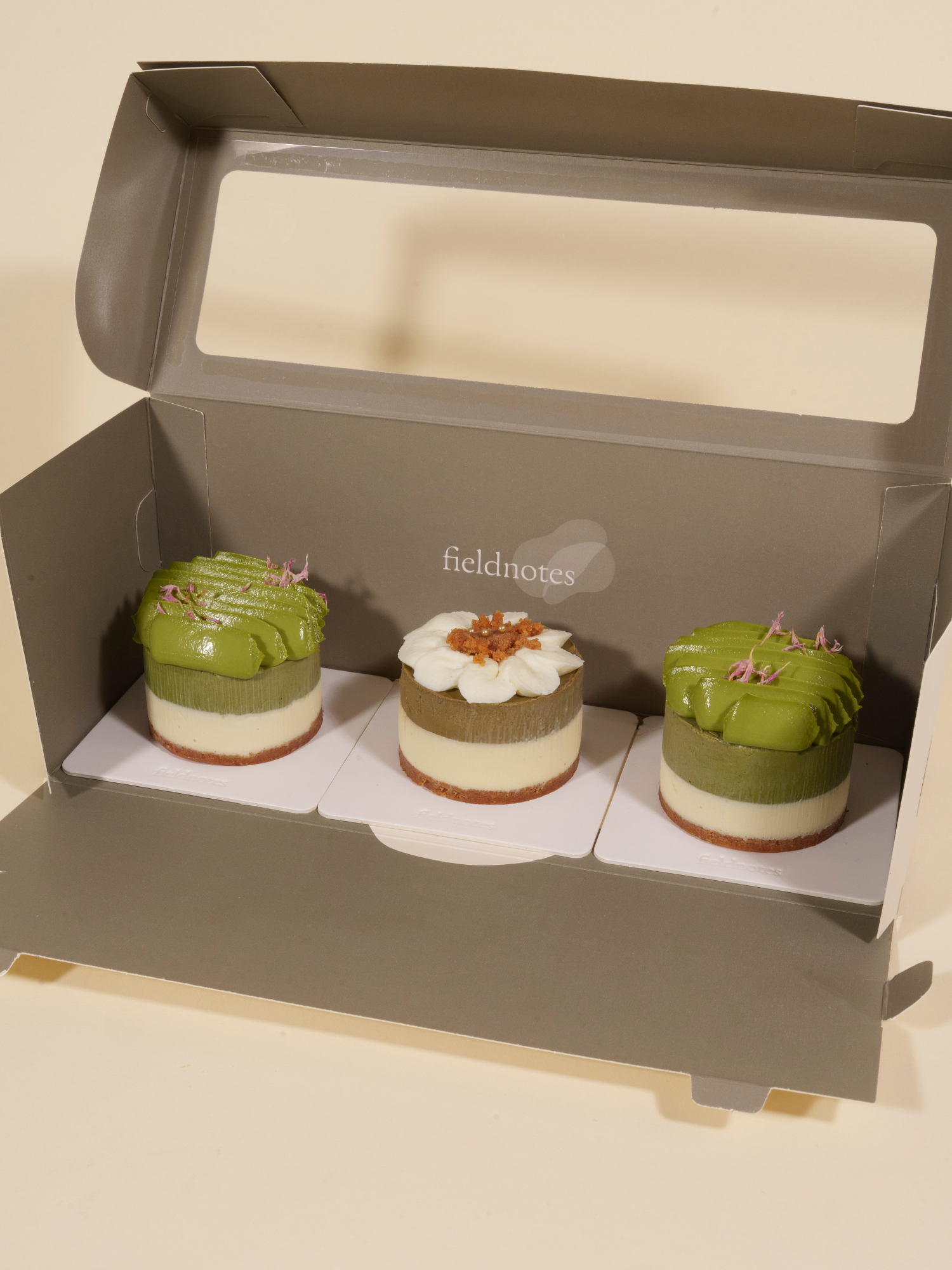 Three small, round birthday cake tarts sit in a partially open gray box; two have green tops and purple garnish, one has a white flower.