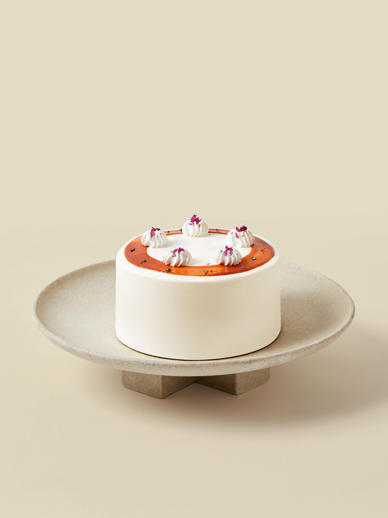 A round Guava Lychee birthday cake by fieldnotes with white frosting, orange glaze, and pink petals sits on a beige tart stand.