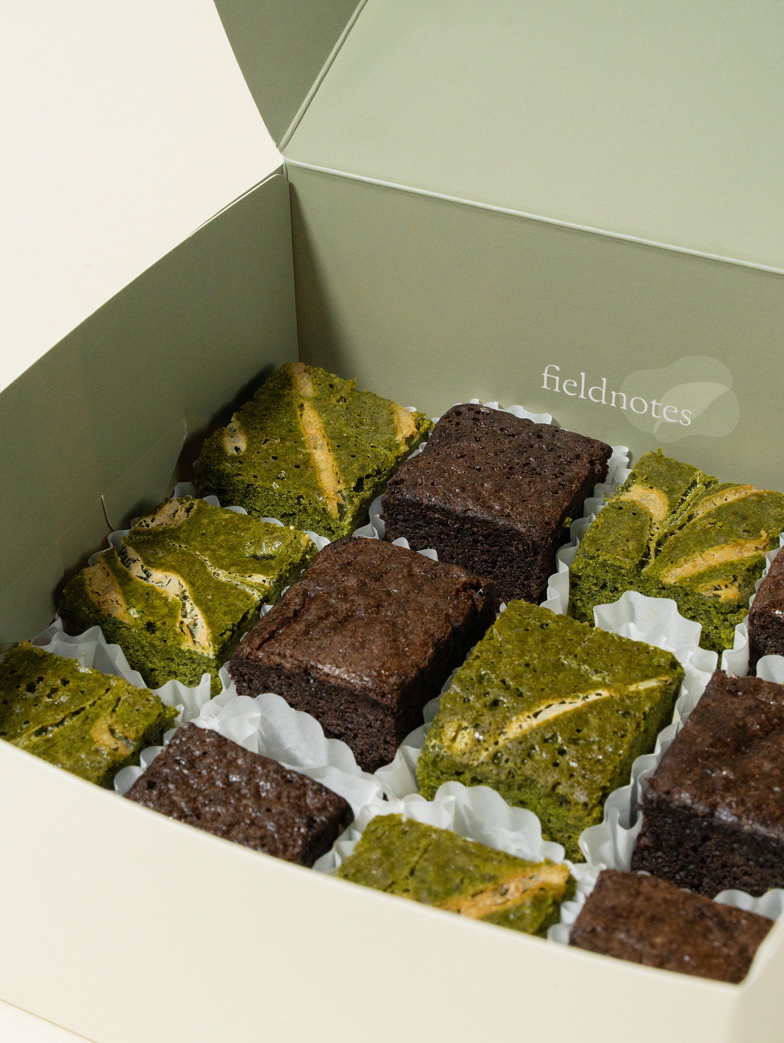 A pastel green fieldnotes box displays brownies and tart treats—matcha white chocolate, chocolate, or birthday cake flavors in paper cups.