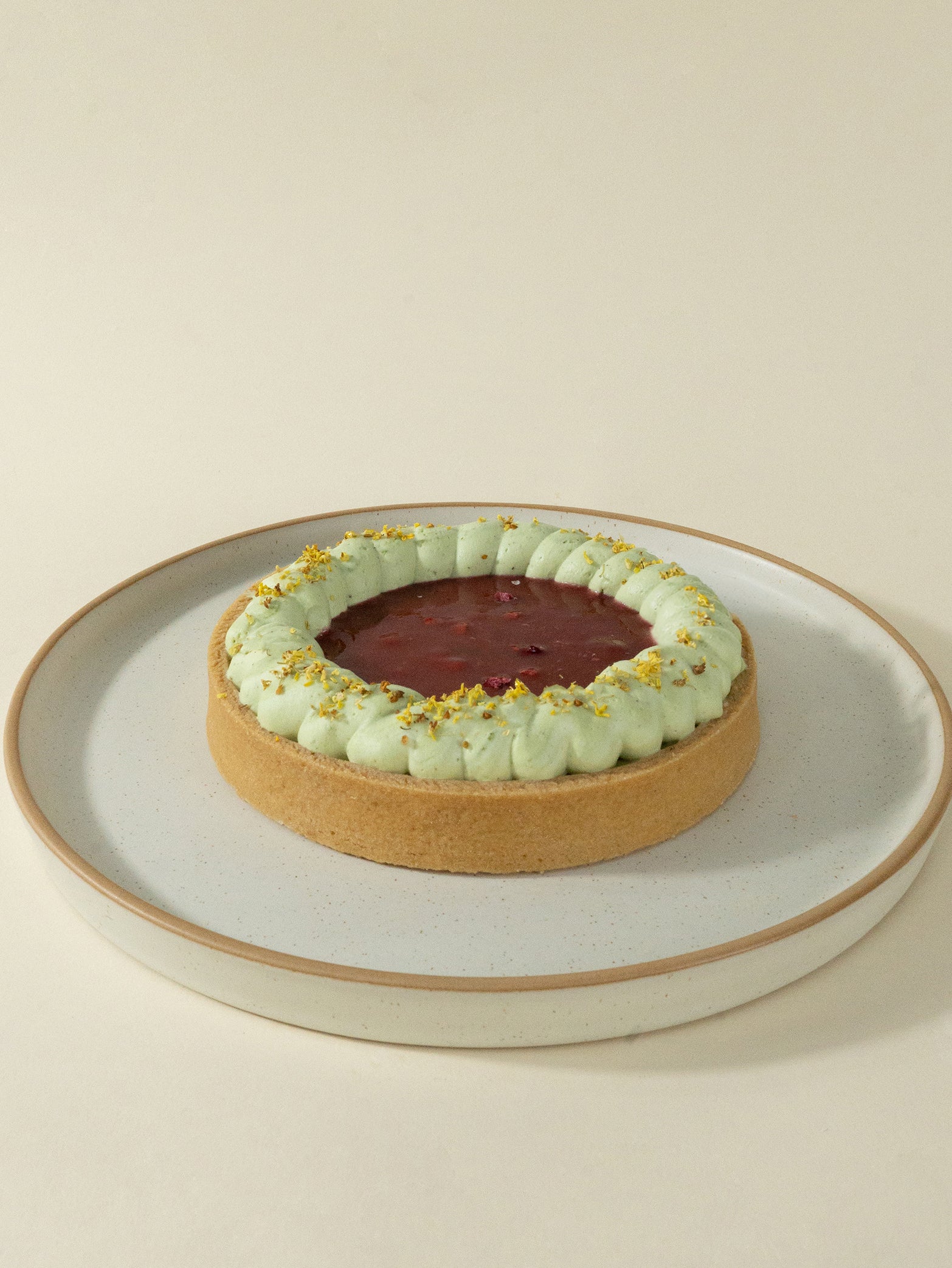 The Raspberry Pistachio Tart features a golden crust, raspberry compote filling, pale green cream, and chopped pistachios, presented on a round white plate against a neutral background.
