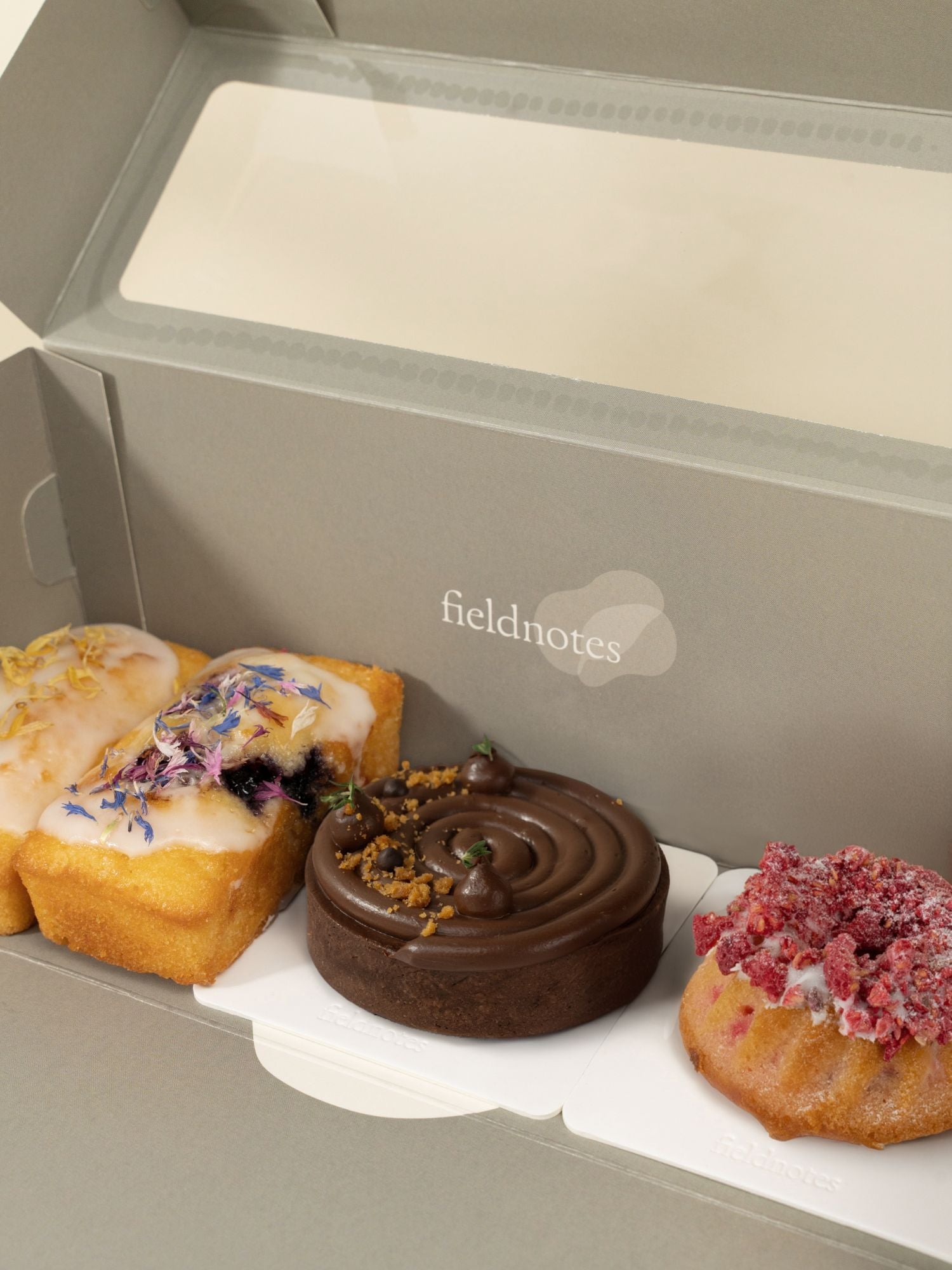The Verdant Mornings | Assorted Bakes Dessert Box features three treats: a rectangular cake with icing and petals, a round chocolate tart, and a small cake with pink crumbs—all in a box labeled fieldnotes.