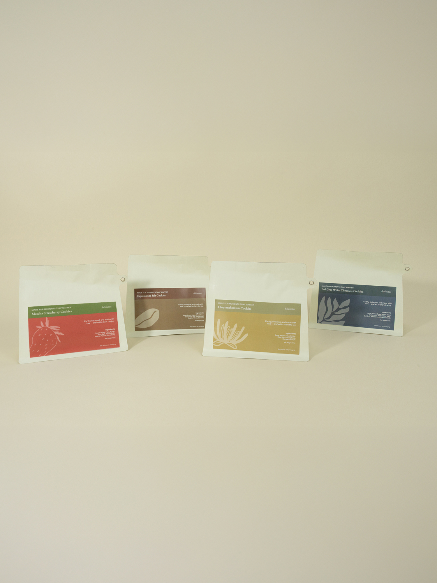 Four white pouches of Nature's Bliss Cookies by fieldnotes stand in a row, each with a colorful label—red, brown, yellow, and blue—showcasing unique flavors like birthday cake or tart, customised with premium ingredients against a plain light background.