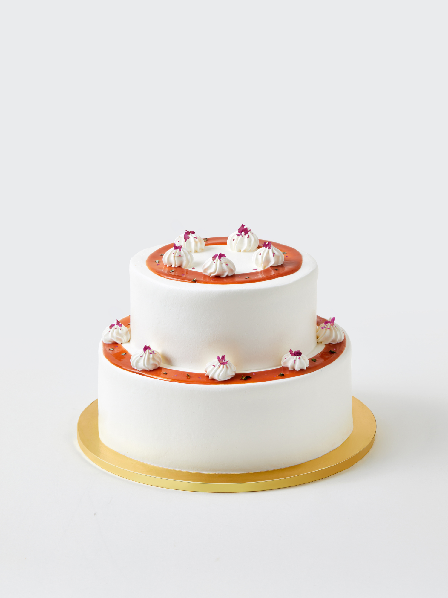 The Guava Lychee Two Tier birthday cake by fieldnotes features smooth white frosting, orange icing rings, and pink guava cream dollops with purple flower toppers, displayed on a round gold cake board against a plain background.