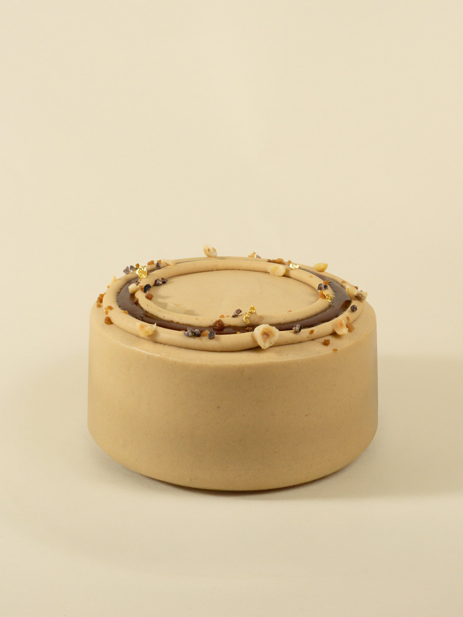 The fieldnotes Hazelnut Praline birthday cake, available for next day delivery, features a round tart-like beige design with smooth frosting, chocolate glaze ring, chopped nuts, gold leaf, and decorative accents—an elegant dessert on a light background.