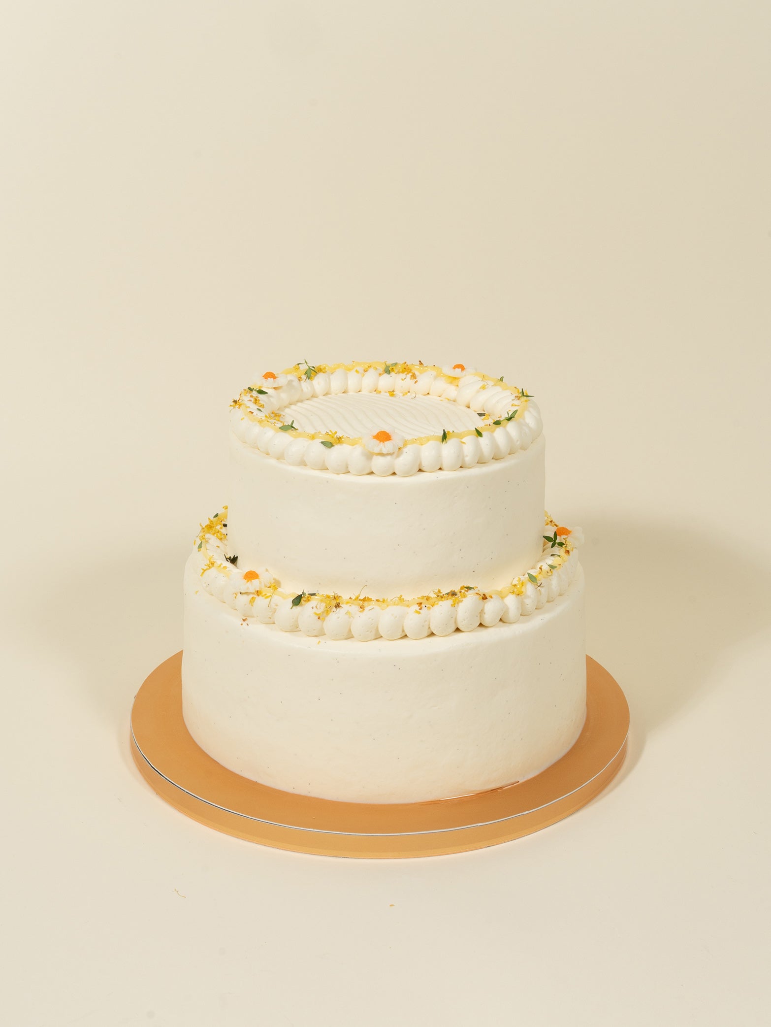 The fieldnotes customised birthday cake is a two-tier tart with smooth white frosting, piped details, yellow and white flowers, and Manuka Honey cream filling on a gold cake board.