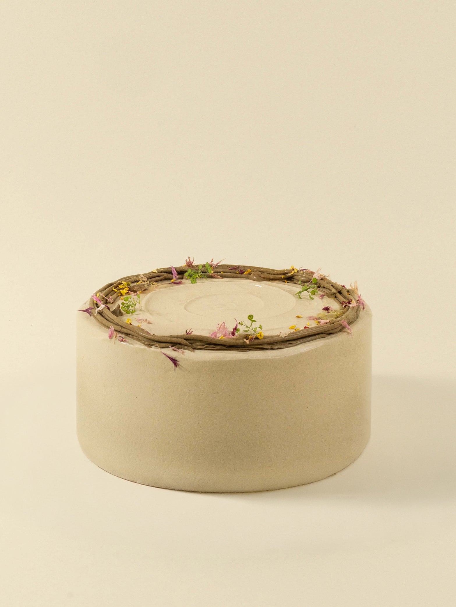 The fieldnotes Oolong Lychee birthday cake is round and minimalist, with a smooth white surface topped like a tart with twigs and small colorful flowers, set against a plain beige background.