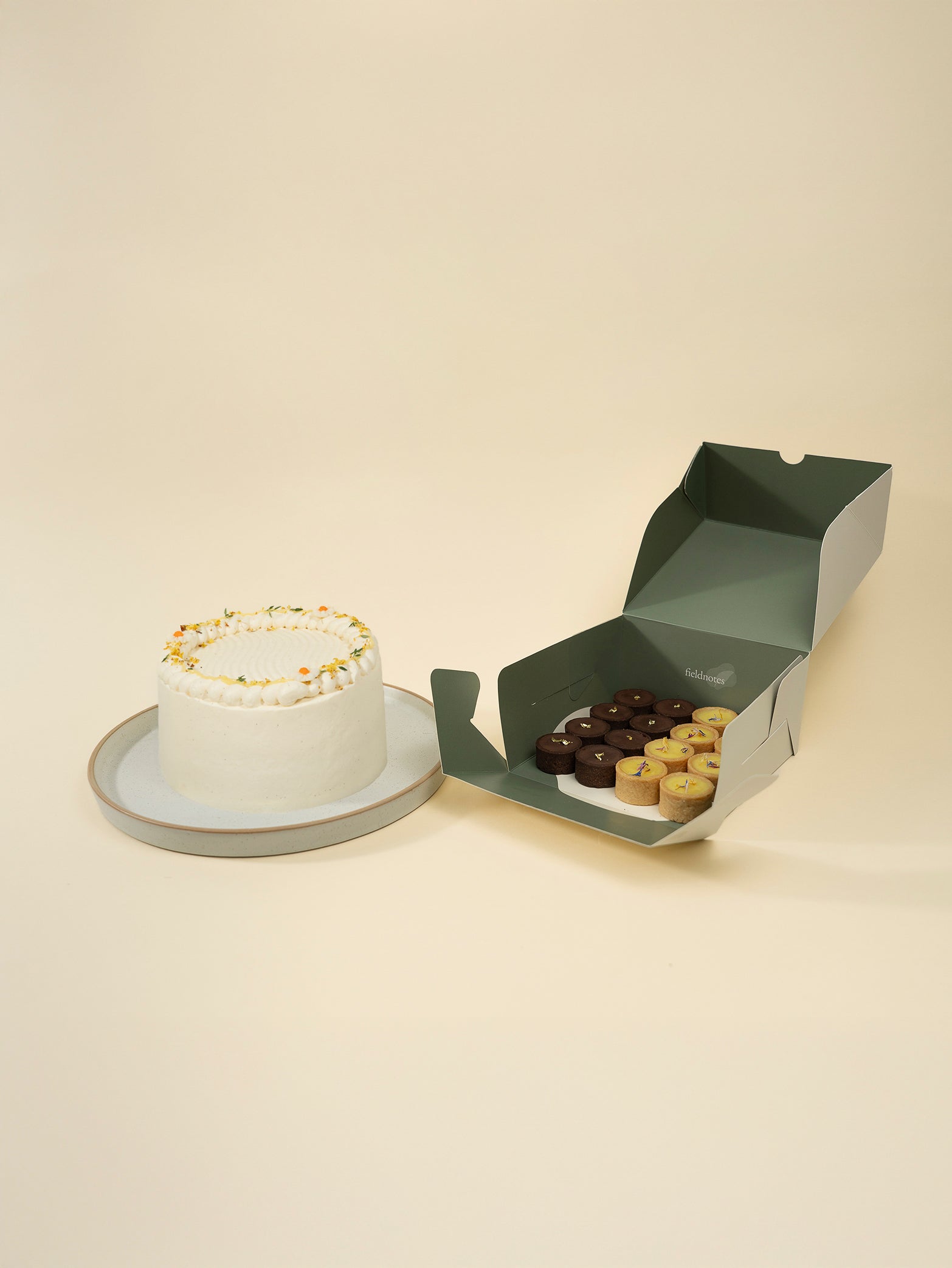 The fieldnotes Towards the Sun Bundle includes a 15CM birthday cake with white frosting and sprinkles, plus a green box of assorted mini cheesecakes and tarts for a delightful celebration.