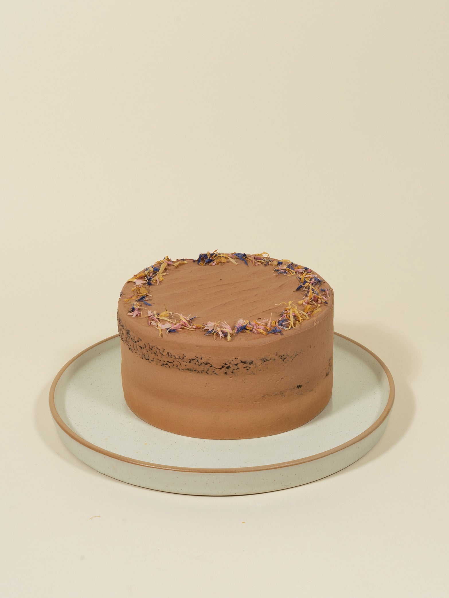 The fieldnotes Vegan Double Chocolate Birthday Cake, a round vegan customised cake with smooth frosting and edible flower ring on top, sits on a white plate with beige rim against a plain background.