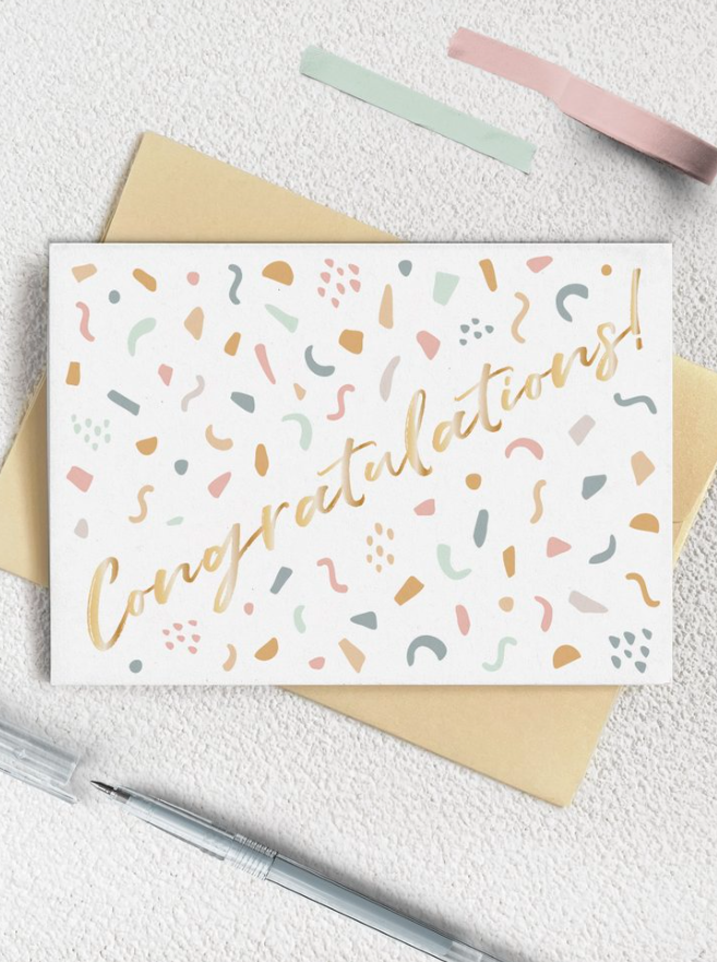 A 384x515 photo of a congratulations card designed with pastel shapes resembling confetti, under the card is a yellow paper, beneath is a clear pen, and above is are two pastel blue and pink tape