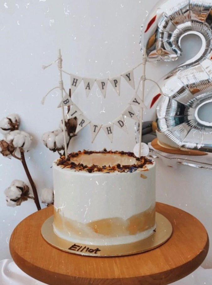A 384x516 photo of a white cake with earthy accents and a handcrafted bunting cake topper that says "Happy Birthday" on a wooden table