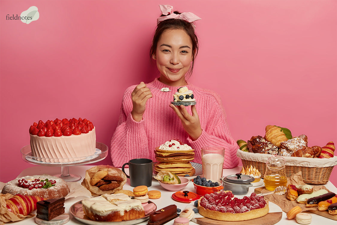 Smiling person in pink holding a cupcake at a table filled with colorful desserts and breakfast items