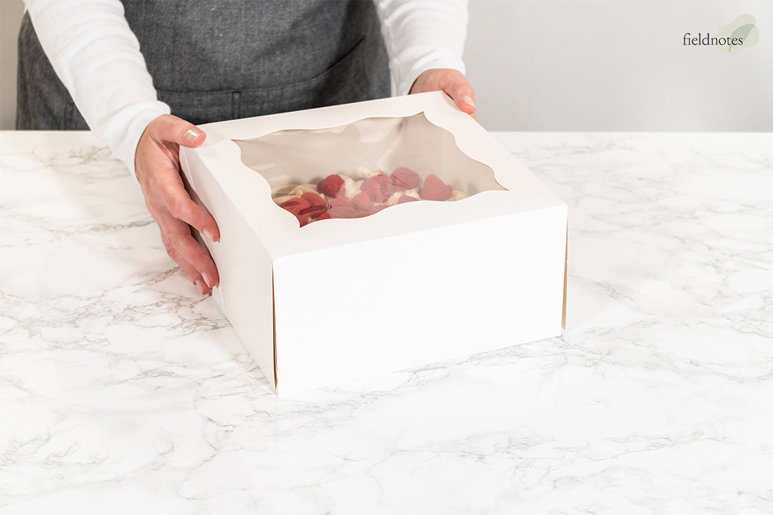 A person holds a white bakery box with a clear window revealing raspberry-topped treats.