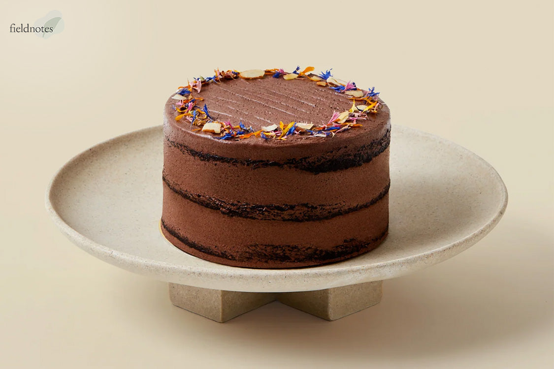 A three-layer chocolate naked cake on a stone pedestal, topped with smooth chocolate frosting, slivered almonds, and colorful dried flower petals.