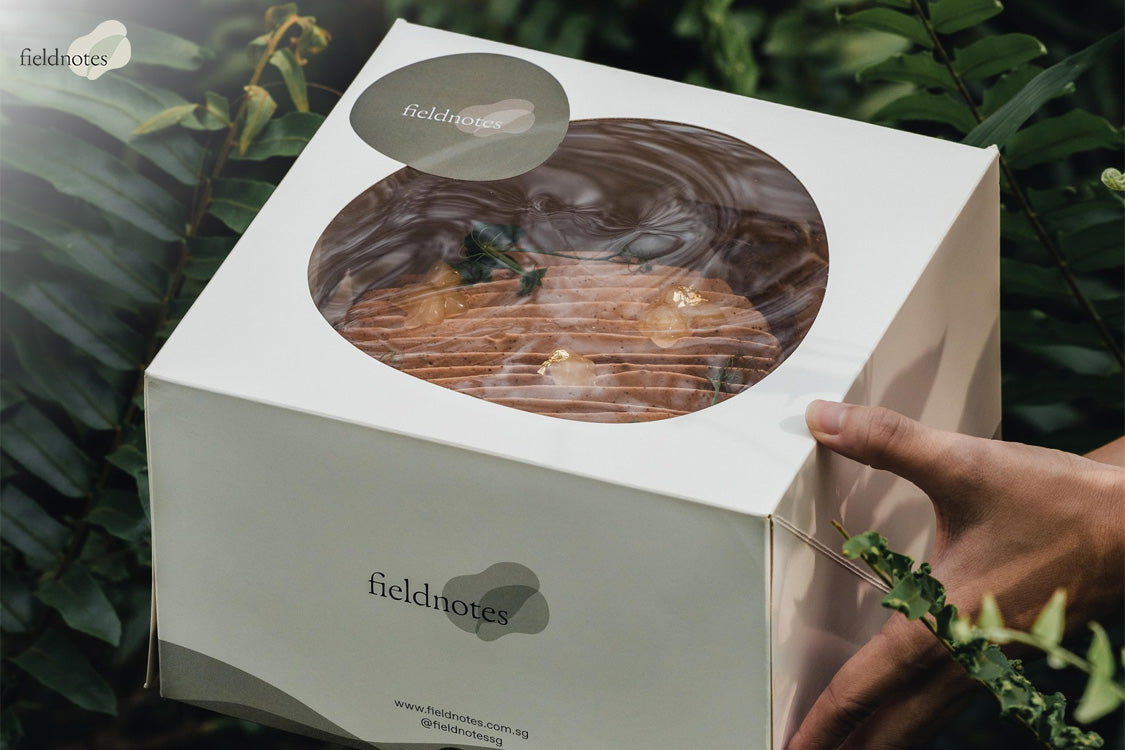 Elegant cake box with circular window showing decorated cake, branded with “fieldnotes” and surrounded by foliage.
