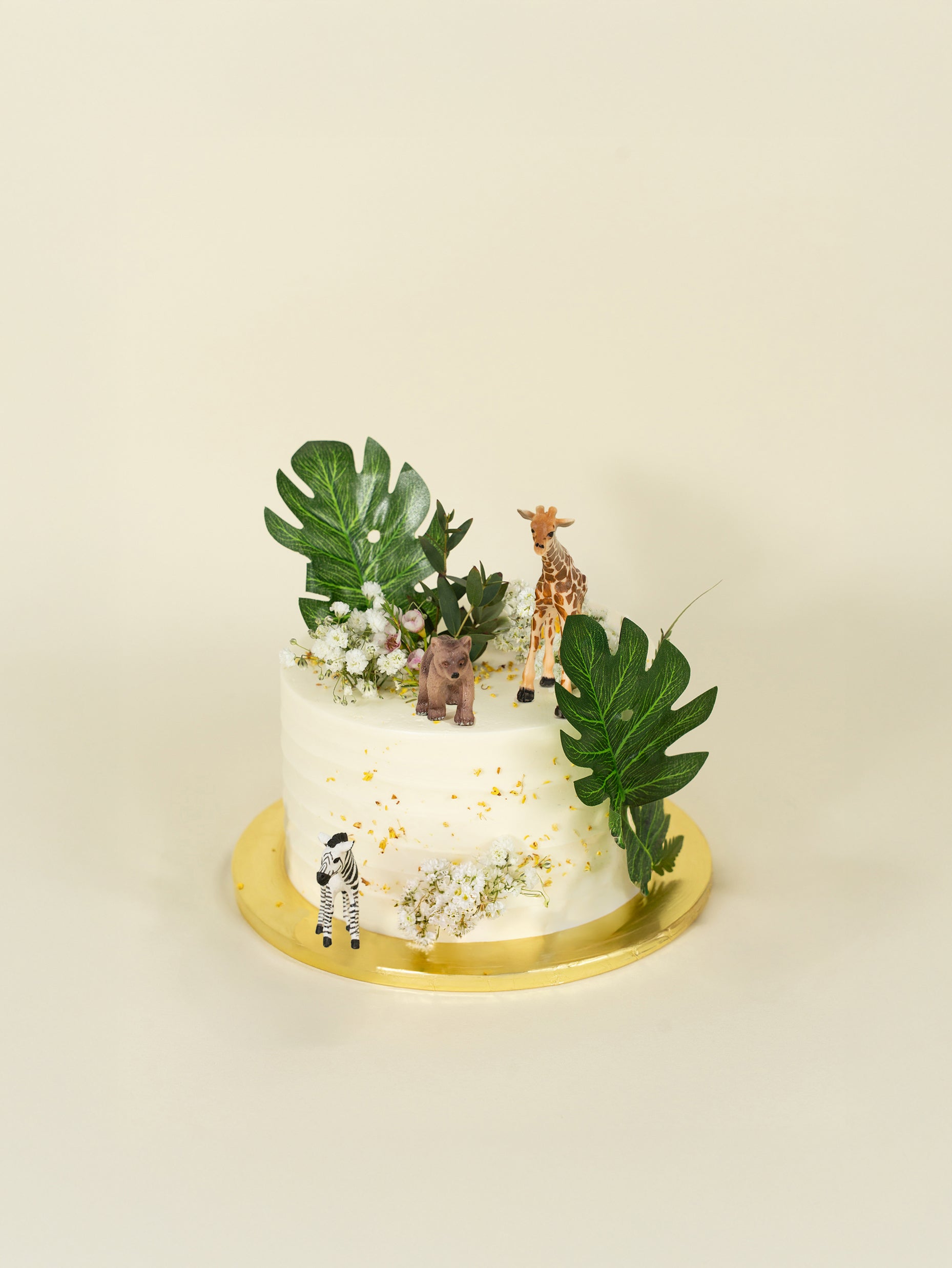 The fieldnotes One tier Rustic Safari birthday cake is a white buttercream tart with green leaves, white flowers, and safari animal figurines.