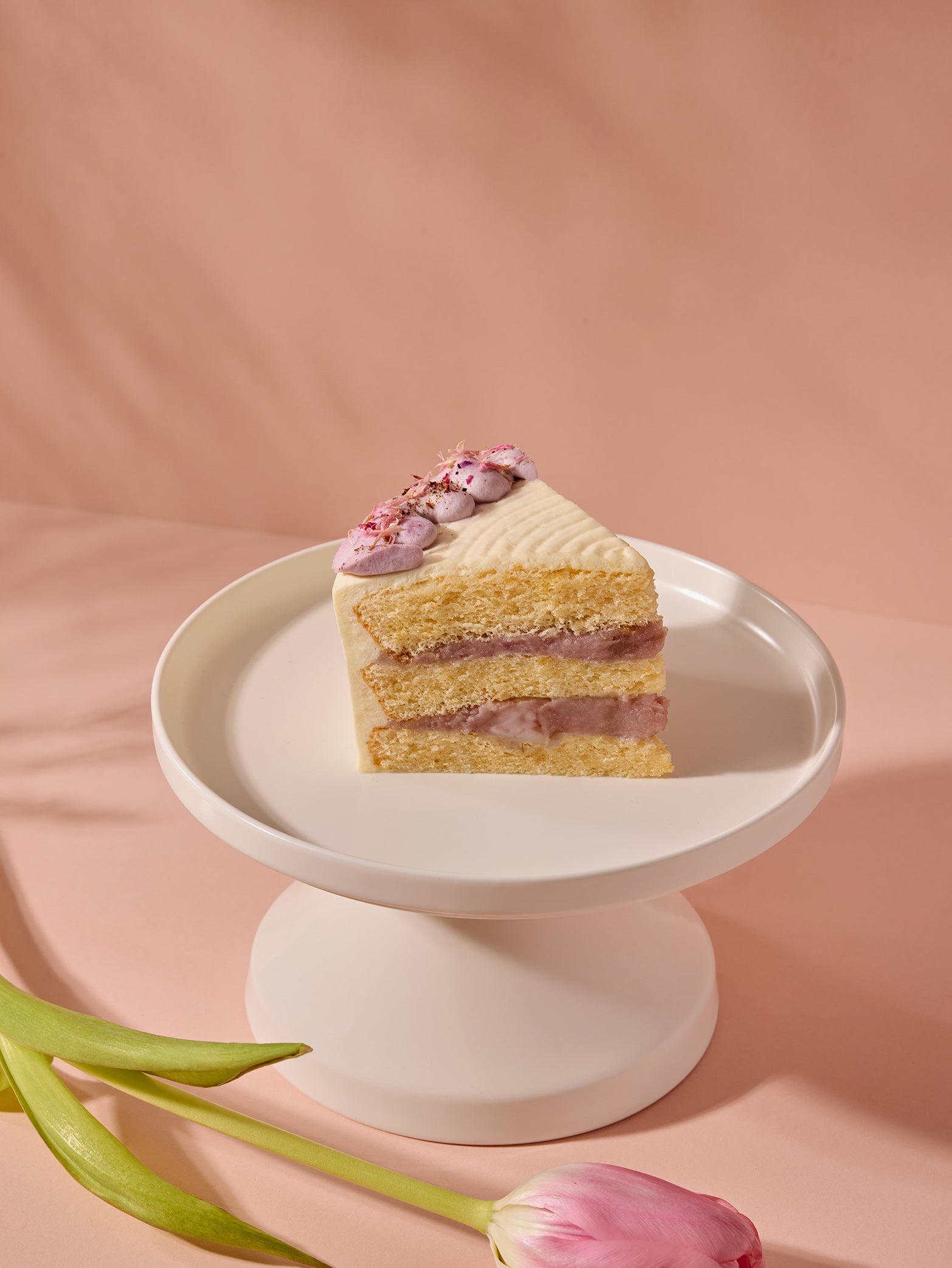 A birthday cake slice of fieldnotes Taro Cake with pink filling and white frosting sits on a stand, tulip beside it on pink.