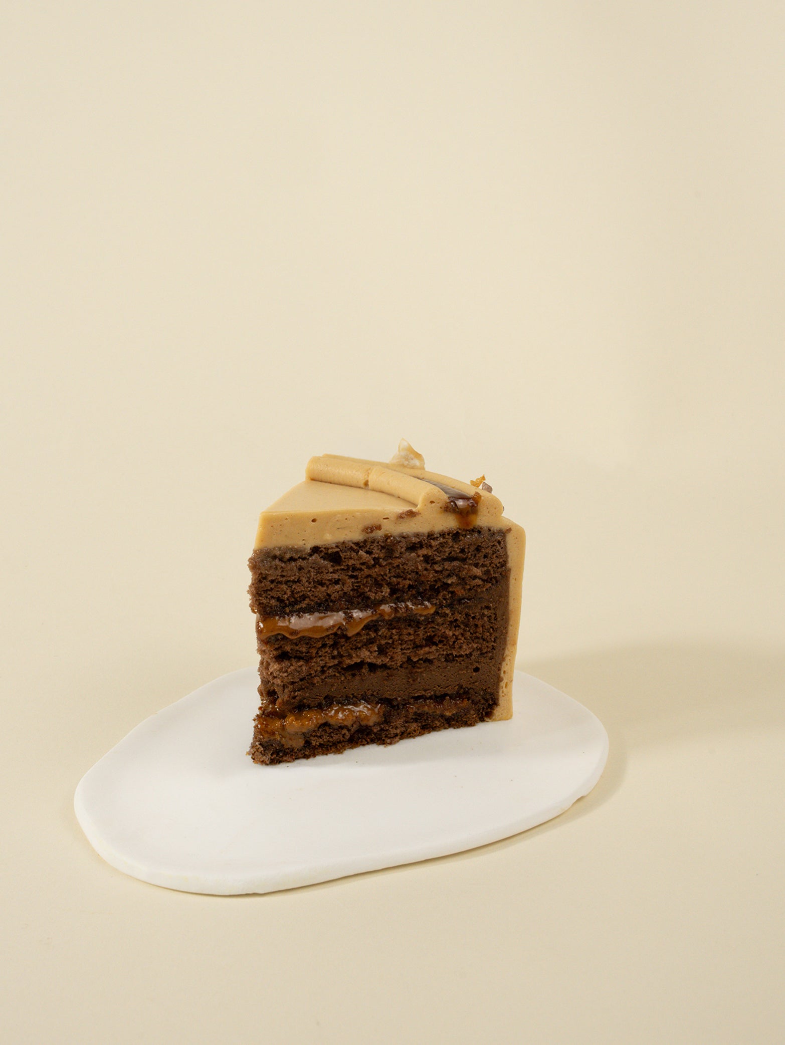 A slice of fieldnotes Hazelnut Praline birthday cake, with light brown hazelnut cream frosting, sits on a white plate. Next Day Delivery.