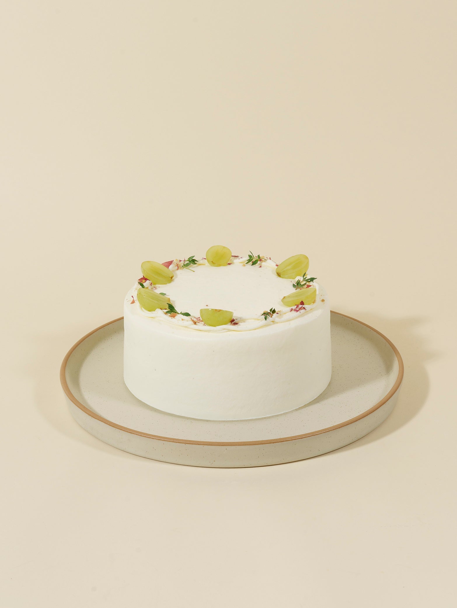The Classic Cakes (Party Size) by fieldnotes is a festive birthday cake, round with white frosting, green grapes, herbs, and flowers on a tart-like base, served on a beige ceramic plate against a light background.