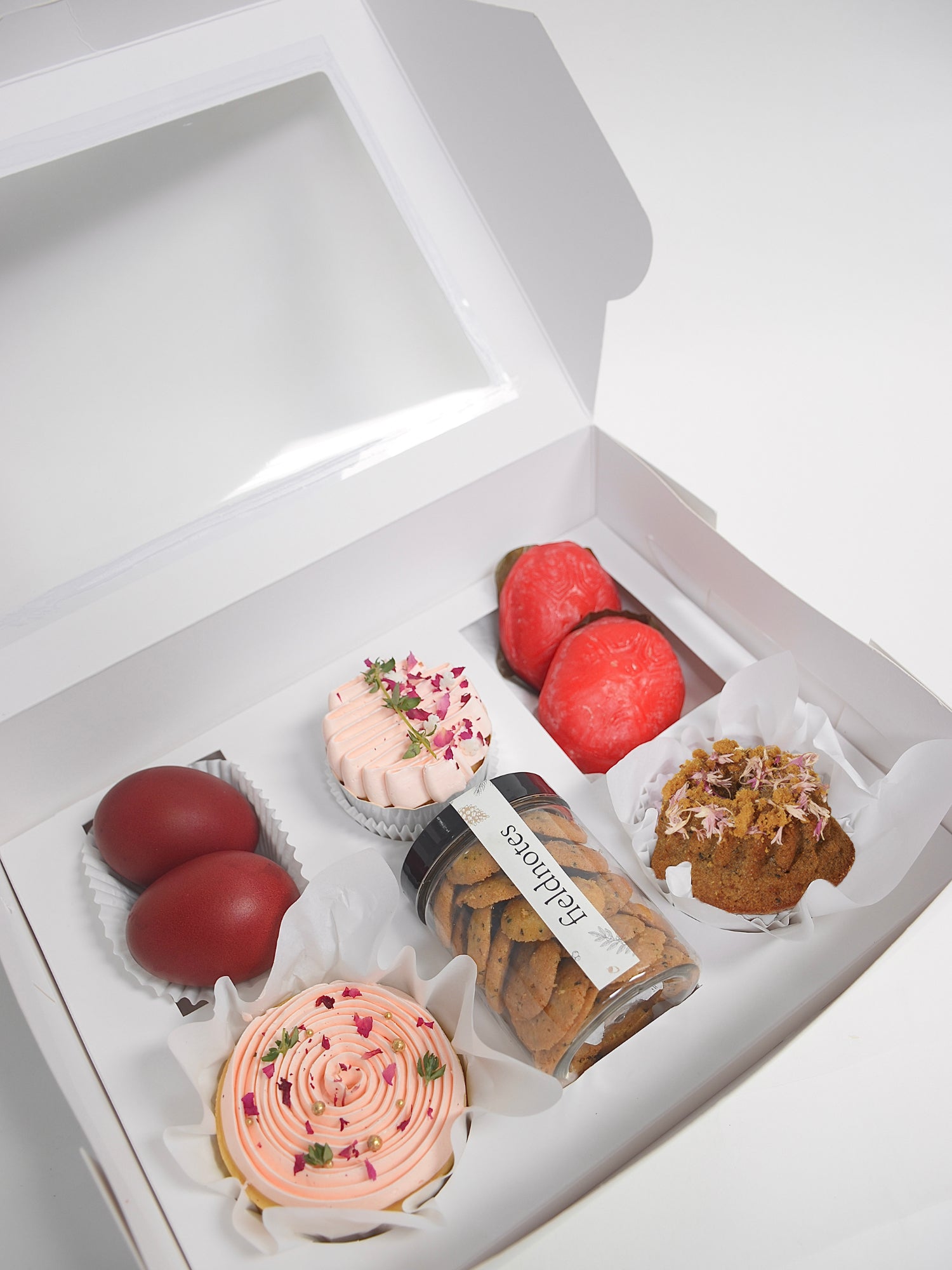 The fieldnotes Stardust Full Month Gift Box Pink has a white box holding red-dyed eggs, cupcakes, a coconut muffin, birthday cake tart, red sweets, and cookies—ideal for baby celebrations.