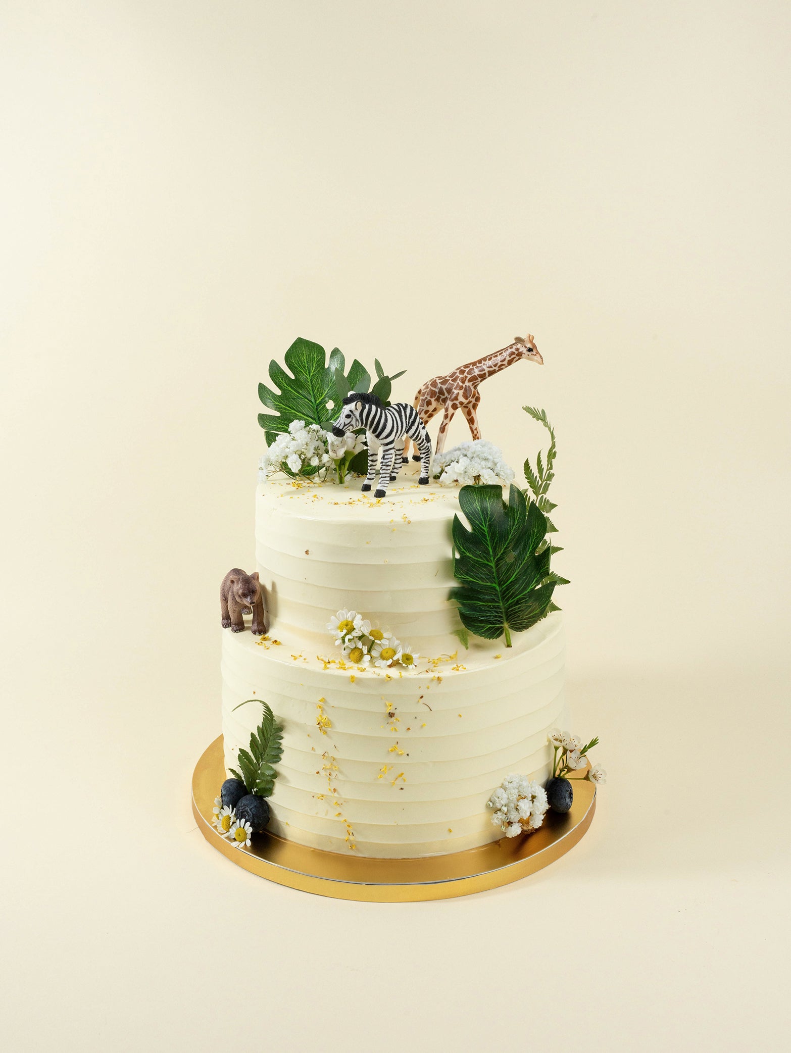 The Rustic Safari two-tier birthday cake boasts a buttercream finish, green leaves, white flowers, and safari figurines on gold board.
