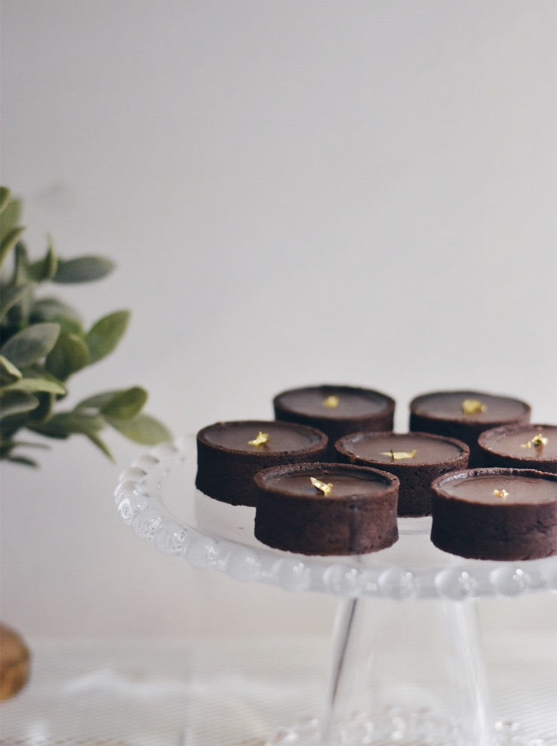 A 384x515 photo of seven chocolate salted caramel tarts arranged in a circle on a glass mount with leaves on the left side of the background